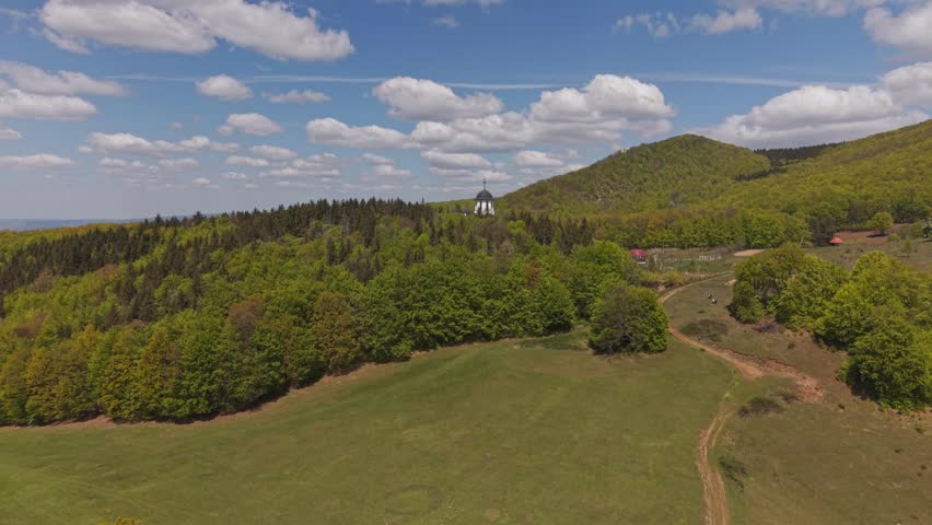 Aerial view of a white church nestled among dense forest in a mountainous landscape under a bright blue sky with scattered clouds.