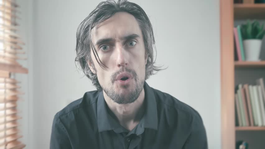 Very surprised man with long hair and beard nodding in agreement while displaying a range of emotions in softly lit setting with wooden blinds and bookshelf background.
