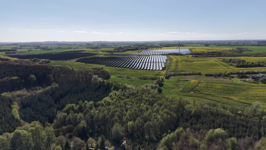 A tracking drone shot revealing the scale of a renewable installation featuring solar arrays and wind turbines amidst blooming agricultural fields in Denmark.