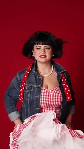 A girl in a beautiful pin-up style dress in a denim jacket with an incredible smile blown by the wind on a dark red background