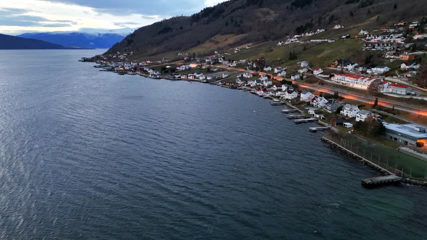 Majestic aerial view flying over a quaint seaside village nestled along the coast of a norwegian fjord, with illuminated houses and roads contrasting against the dark, rippling water at dusk