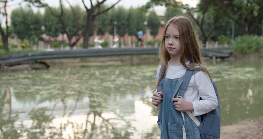 Portrait of pretty little girl with schoolbag stands near pond. Cute elementary student smiles in green urban park. Knowledge for all children program