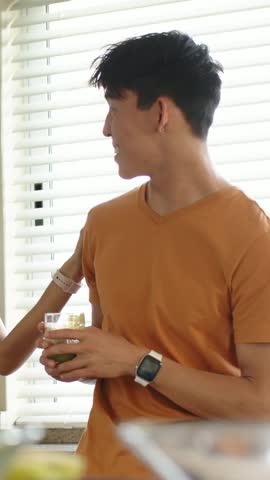 Vertical video: Smiling man in orange tee with glass, sampling green wedge woman handing at sink. Kitchen, couple, beverage, tasting, daylight, countertop, vertical video