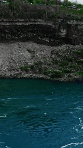 Deep blue river against cliffside, rugged limestone walls with visible strata, swirling currents and foam, pebbled shoreline and sparse vegetation on clifftop park, overcast sky creating moody