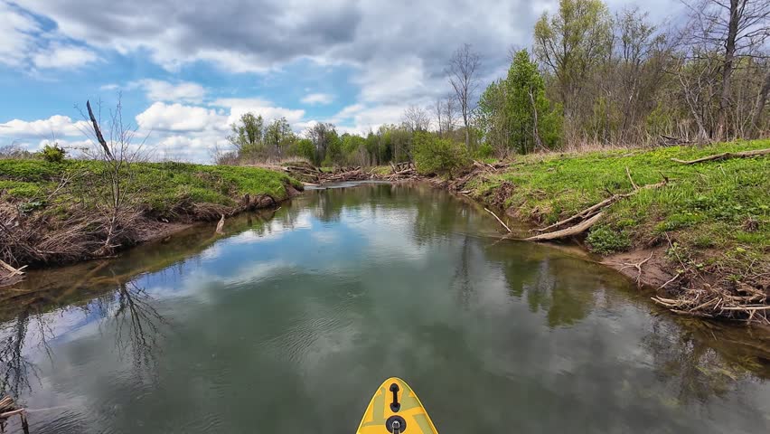 Wide Creek Mirror Surface Under Sky, Yellow Bow Visible, Gentle Meander Between Grassy Banks, Reflective Water, Scattered Driftwood And Spring Greenery Invite Calm Voyage