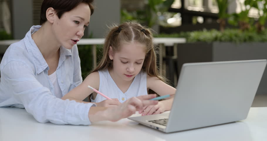 Portrait of mother and little daughter work on assignment at laptop in outdoor office. Woman teacher explains task to junior schoolgirl at computer on terrace