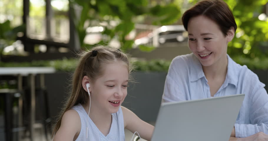 Portrait of happy little girl listens to video lesson via laptop as mother supports nearby in cafe. Woman sits near smart schoolgirl learning online in office