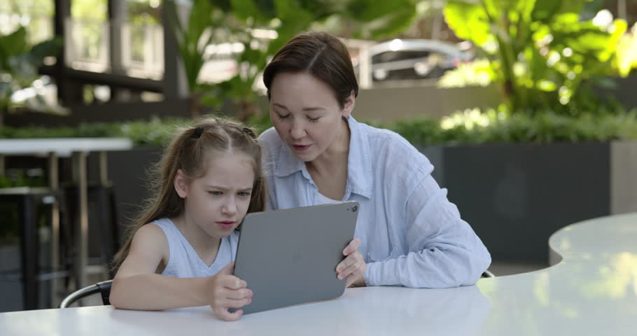 Portrait of caring mother with little daughter uses digital tablet as sits in cafe. Woman and lovely girl watch online video via mobile device on city street