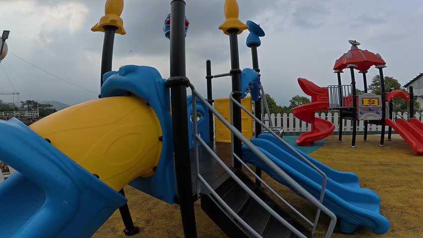 Wide angle view of colorful playground equipment featuring blue and yellow slides, stairs, and play structures in an outdoor public park. Modern children’s playground with bright plastic slides under cloudy sky, ideal for family recreation, childhood activities, education, kindergarten, and leisure concepts.