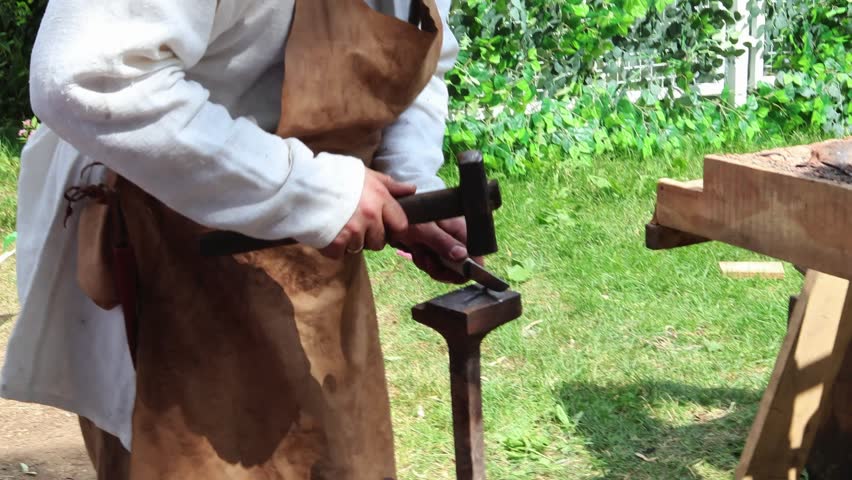 Blacksmith in traditional attire wields hammer over an anvil outdoors, demonstrating timeless art of blacksmithing, historical craftsmanship and manual labor.