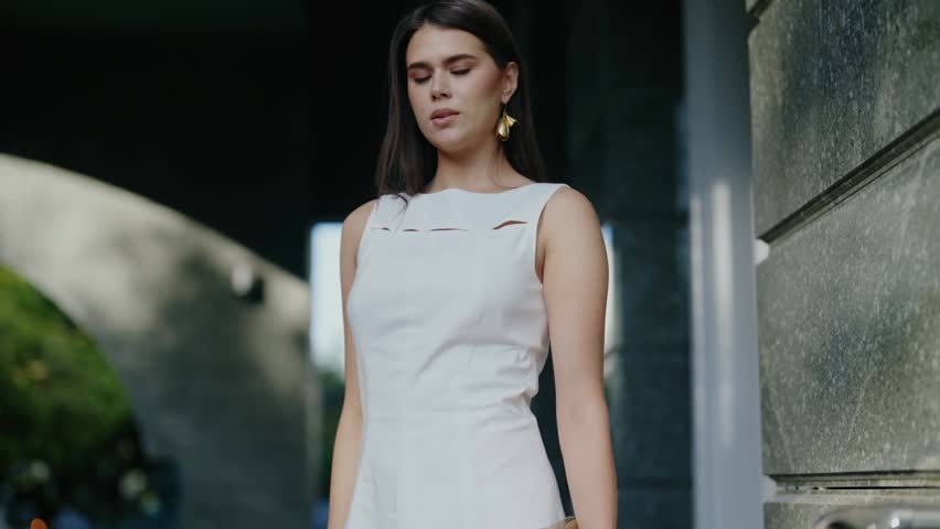 A young woman in a stylish white dress poses near a building. She exudes confidence and elegance, capturing a sense of urban chic. Golden earrings add a touch of glamour to her look.