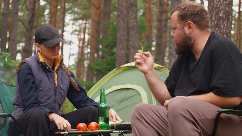Happy young couple sitting on camping chairs in woods, talking and laughing together, grilling meat on barbecue picnic during their trip to the fresh air and drinking beer
