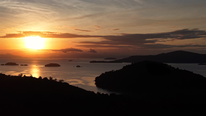 A wide panoramic view of a vibrant sunset over a calm tropical bay with reflections on the water and island silhouettes