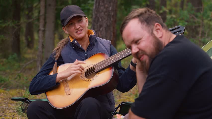 Young smiling woman playing an acoustic guitar and singing songs for a bearded man while sitting in camping chairs in the woods. Happy friends enjoying their vacation and music together in nature