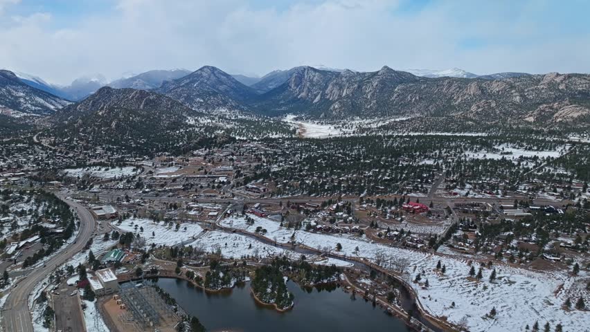 Estes Park town with lake shoreline and snow covered Rocky Mountains under cloudy sky, aerial drone overview