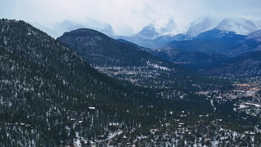 Estes Park forested valley with snow covered Rocky Mountains and cloudy sky, aerial drone overview