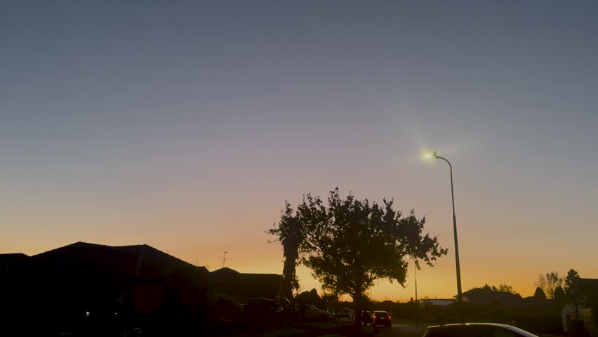 Golden sunset over a quiet suburban neighborhood in Auckland, New Zealand, featuring silhouetted houses, trees, parked cars, and a glowing streetlight beneath a deep blue evening sky. A calm residential atmosphere captures the peaceful transition from day to night.
