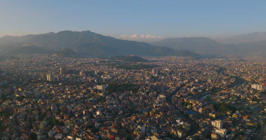 Aerial shot of overlooking unmanaged urban city Kathmandu, Nepal, surrounding hills and distant Himalayan peaks illuminated by warm evening golden hour light.
