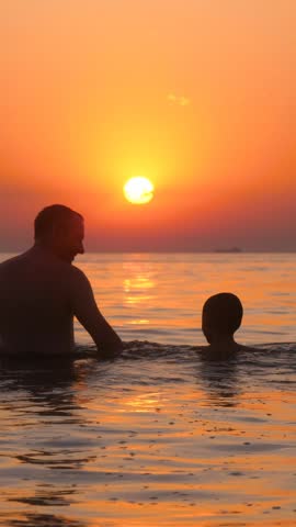 Father and son enjoying sunset in water. Dad and child share joyful moments and laughter at beach. Warm atmosphere creates memorable family bonding experience. Vertical shot captures beautiful scenery