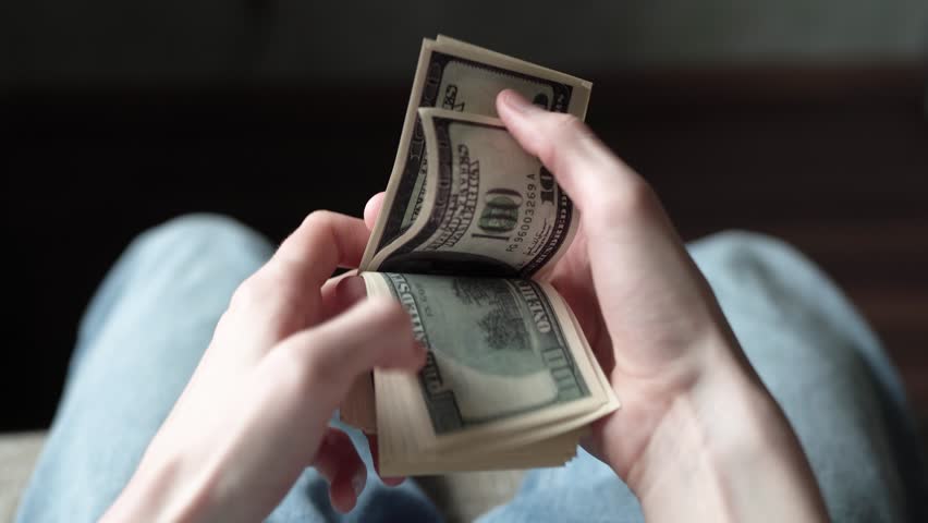 A young person counts a stack of cash. The hands of the individual are focused on the bills, emphasizing the act of handling money