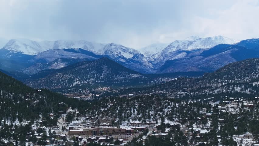 Estes Park mountain panorama with forest valley and snow covered peaks beneath cloudy sky, aerial drone overview