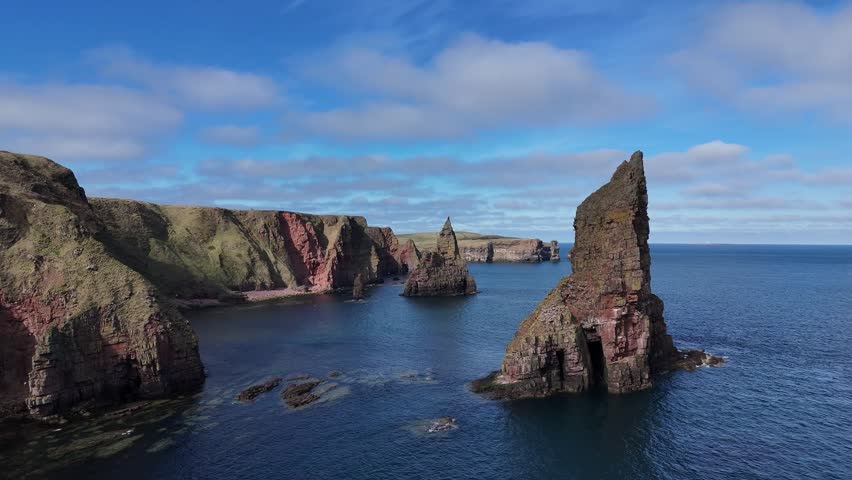 Scotland - A cinematic flight through the incredible Duncansby Sea Stacks on a bright, sunny day with natural cloud. Calm and turquiose sea water with nesting birds surrounding the area. 