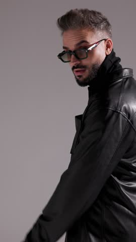 Stylish man in sunglasses and leather jacket is fixing his collar, looking around, and crossing his arms on grey studio background.