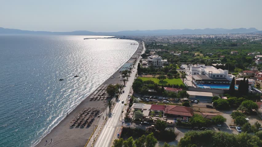 Aerial drone footage of Kalamata Bay on the Peloponnese coast of Greece, showing a long beachfront road running beside a pebble beach and the sparkling Mediterranean sea. The scene includes organized beach umbrellas, seaside hotels, villas, palm trees, green gardens, parked cars, small boats offshore, distant city buildings and hazy mountain silhouettes across the bay. This scenic daytime coastal view is suitable for travel advertising, tourism promotion, Mediterranean vacation content, beach resort projects, summer lifestyle videos, coastal city overviews, hotel and hospitality themes, seaside real estate, urban infrastructure and documentary footage about Greece.