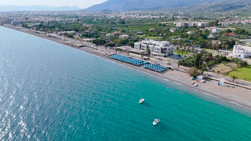 Aerial drone footage of Kalamata beach resort on the Peloponnese coast of Greece, showing two small boats floating in clear turquoise Mediterranean water near a pebble shoreline. The scene includes organized beach umbrellas and loungers, seaside hotels, a coastal road, villas, palm trees, green hills, olive groves and scattered residential buildings on the slopes. This bright summer coastal view is suitable for travel advertising, tourism promotion, Mediterranean vacation content, beach resort projects, boating and nautical themes, hotel and hospitality videos, seaside lifestyle, coastal real estate and documentary footage about Greece.