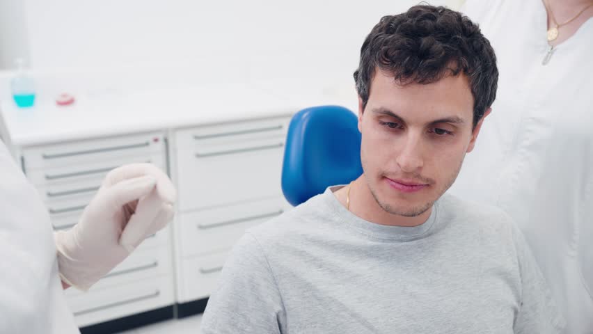Young man sitting in a dental chair and trying on a new pair of transparent aligners. An orthodontist and her assistant are helping him