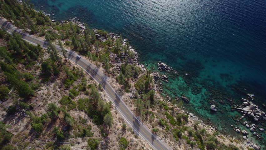 Drone shot of Lake Tahoe with detailed water textures, rocky coast, and forest-lined road in alpine setting.