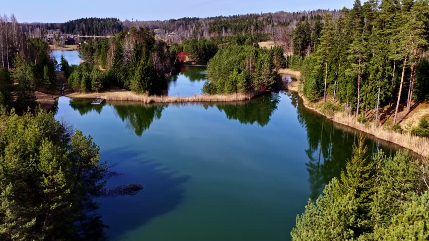 Aerial view glides above a forested lake with small dock and distant houses visible