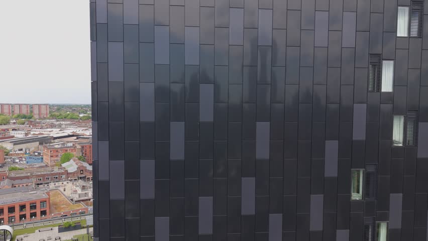 Cinematic drone footage rising vertically alongside a modern black high rise building in Birmingham UK. Urban skyline and city landscape visible in the background during the smooth aerial ascent.