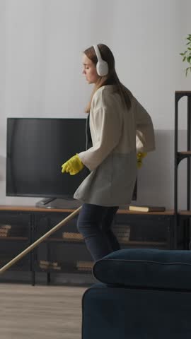 Young woman wearing headphones and rubber gloves enthusiastically mopping the living room floor while listening to music near television and modern home decor