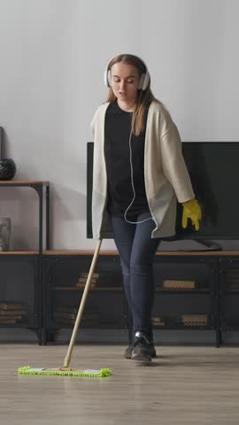 Young woman wearing headphones and rubber gloves mopping wooden floor in modern living room interior, listening to music while doing relaxed household cleaning