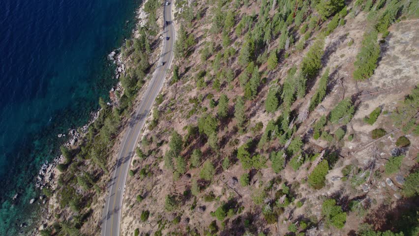 Drone footage showing a winding road along a blue alpine lake with vehicles traveling through a natural landscape.