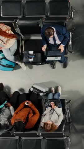 Vertical video Boarding flight, departing airplane and tourists in airport lounge standing up from chairs, going to aircraft bridge. Going to boarding gate, trolleys and passengers leaving airport