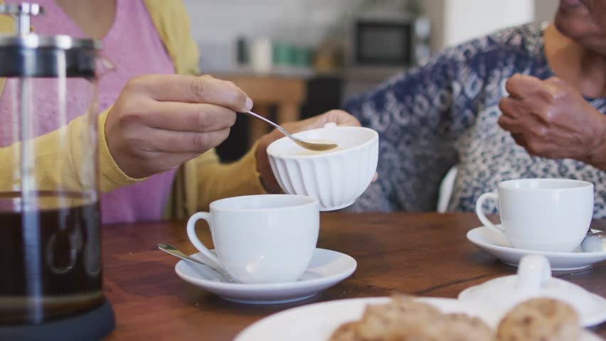 Daughter spooning sugar into cup offering mom care in kitchen, tech icons drifting over hands, cups. Intergenerational, familybond, tea, coffee, dining, porcelain, table