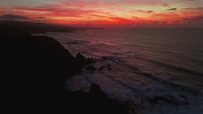 Deep red sunset over ocean waves and dark rocky coastal cliffs.