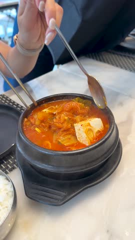 A person uses a spoon and chopsticks to eat spicy Korean kimchi stew from dolsot