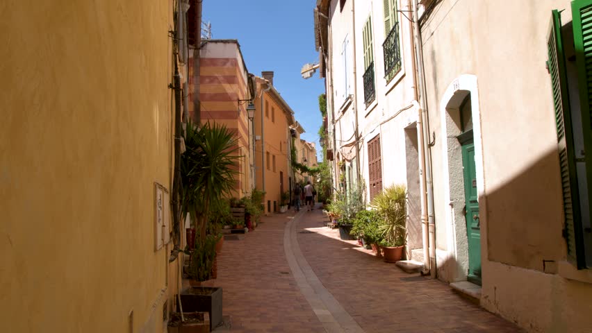 Static wide shot of a sunny European street with potted plants and shuttered windows