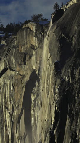 Vertical Screen: Yosemite National Park Firefall phenomenon with water streaming down massive granite rock face, sunlight illuminating the cliff with pine trees silhouetted against blue sky