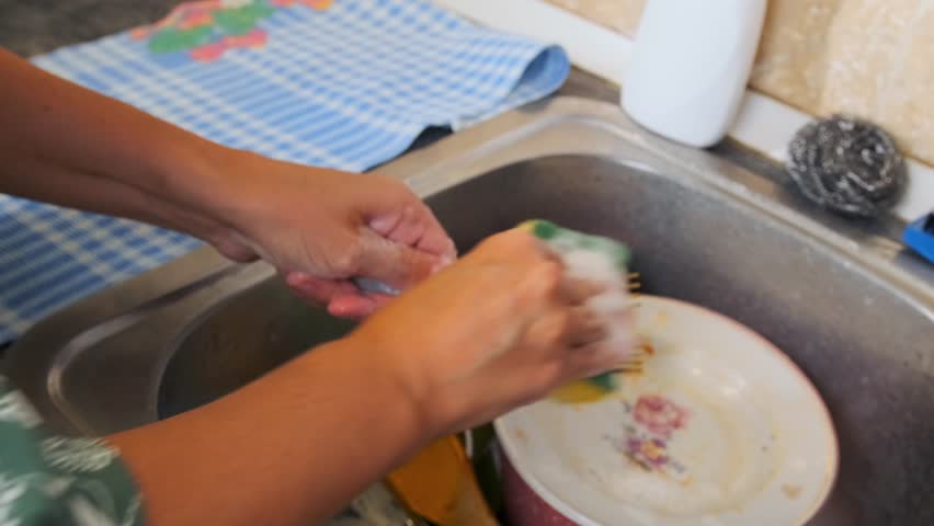 Side view of woman washing dirty dishes in kitchen sink using sponge and detergent. Everyday home routine, dishwashing process, cleaning cookware, domestic chores and hygiene concept.