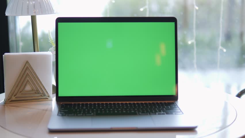 Laptop with green screen sits on white table near window. Hand places cup beside laptop. Background shows natural light and blurred greenery. Minimalist decor includes geometric candle holder