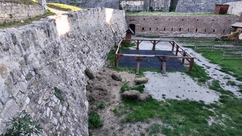 Visitors can see the stone walls of an old fortress. There are grassy areas and a wooden structure in the courtyard. The sun is shining on the ancient stones as nature surrounds the site.