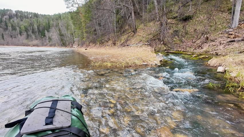 Clear mountain stream along riverbank, pebbled shoal and mossy pine slope. Kayak bow visible as viewpoint, crystal water flowing over stones, cool overcast light, remote wilderness camping