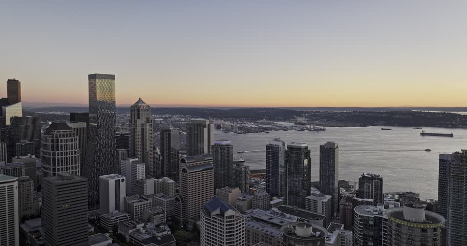 Slow aerial drone footage captures downtown Seattle cityscape at sunset, showcasing skyscrapers reflecting golden hour light, with ferry traffic on Puget Sound and Olympic Mountains in background.
