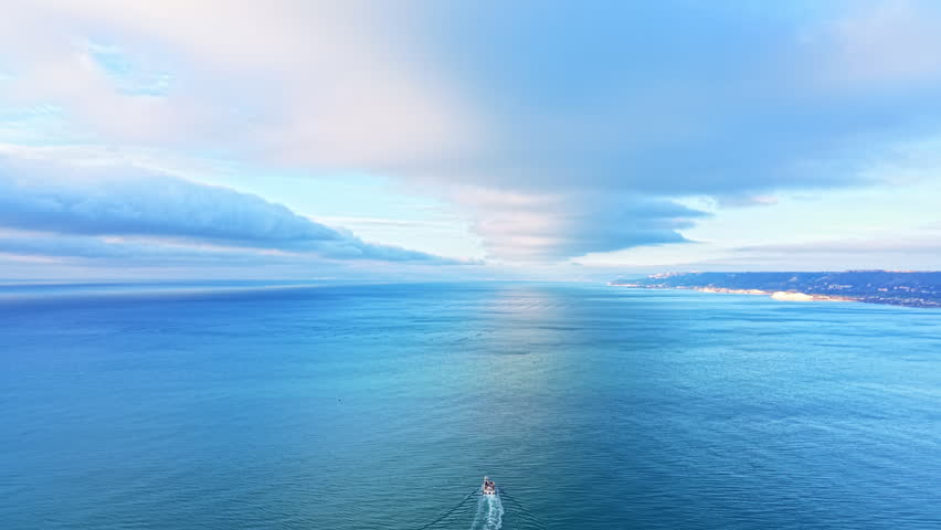 A boat moves through the blue ocean water with waves behind it. Clouds are present in the sky, and land is visible in the distance.