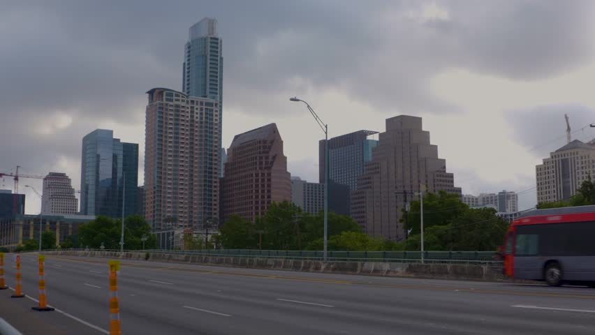 Austin skyline view from highway bridge with cars and buses moving. Tall city buildings under cloudy sky. Urban transportation scene.