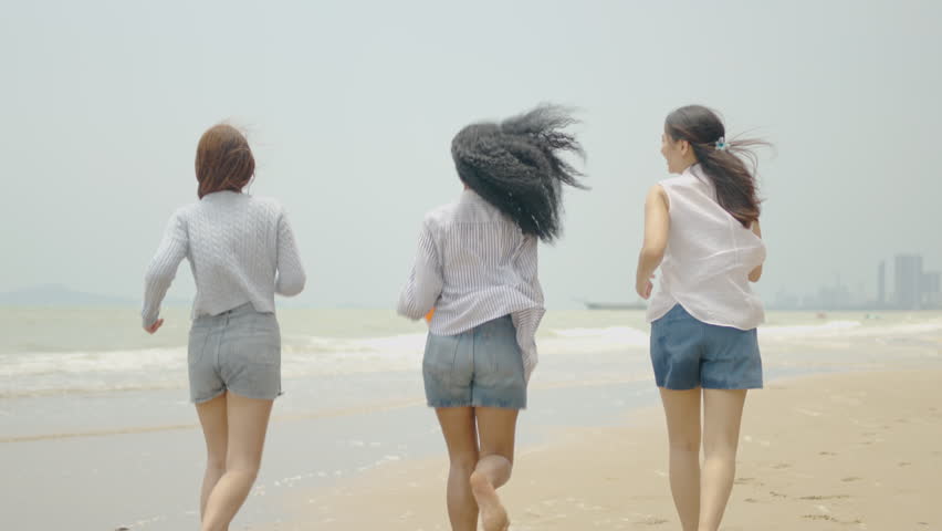 Three young diverse multiethnic female friends running and jumping together on a sandy beach holding a colorful inflatable beach ball, enjoying their joyful summer vacation holiday from behind.